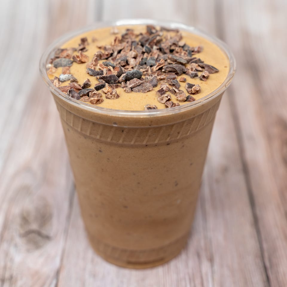 Creamy brown smoothie topped with cacao nibs in a clear plastic cup on a wooden table.