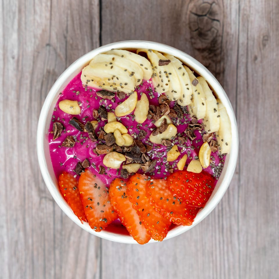 Overhead view of a vibrant purple smoothie bowl topped with banana, strawberries, cashews, cacao nibs, and chia seeds.