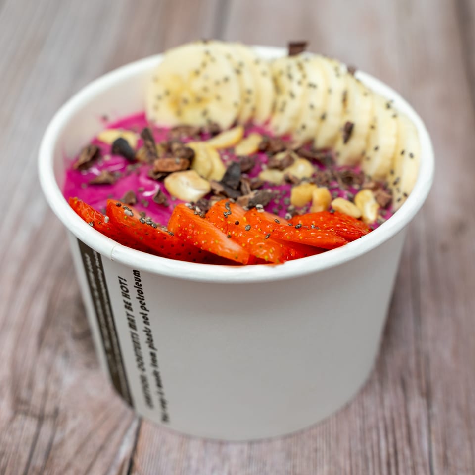 Acai bowl topped with sliced banana, strawberries, nuts, and chia seeds in a paper cup on a wooden table.