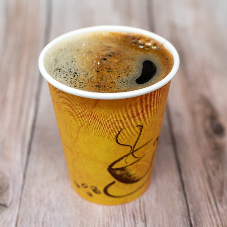 Foamy black coffee fills a yellow illustrated takeaway cup resting on a wooden table.