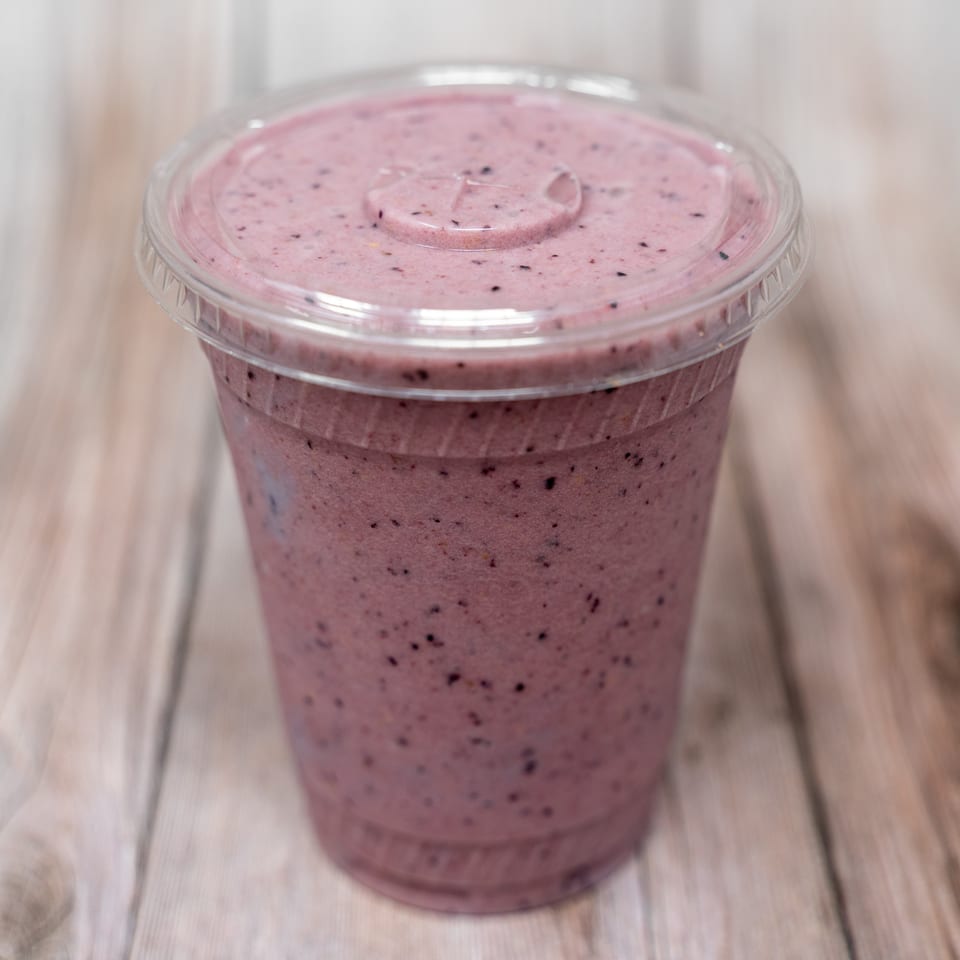 Purple berry smoothie in a clear plastic cup with a lid, resting on a wooden table.