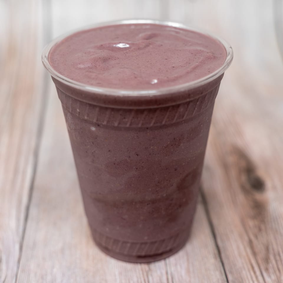A clear plastic cup holds a thick purple berry smoothie on a light wooden table.