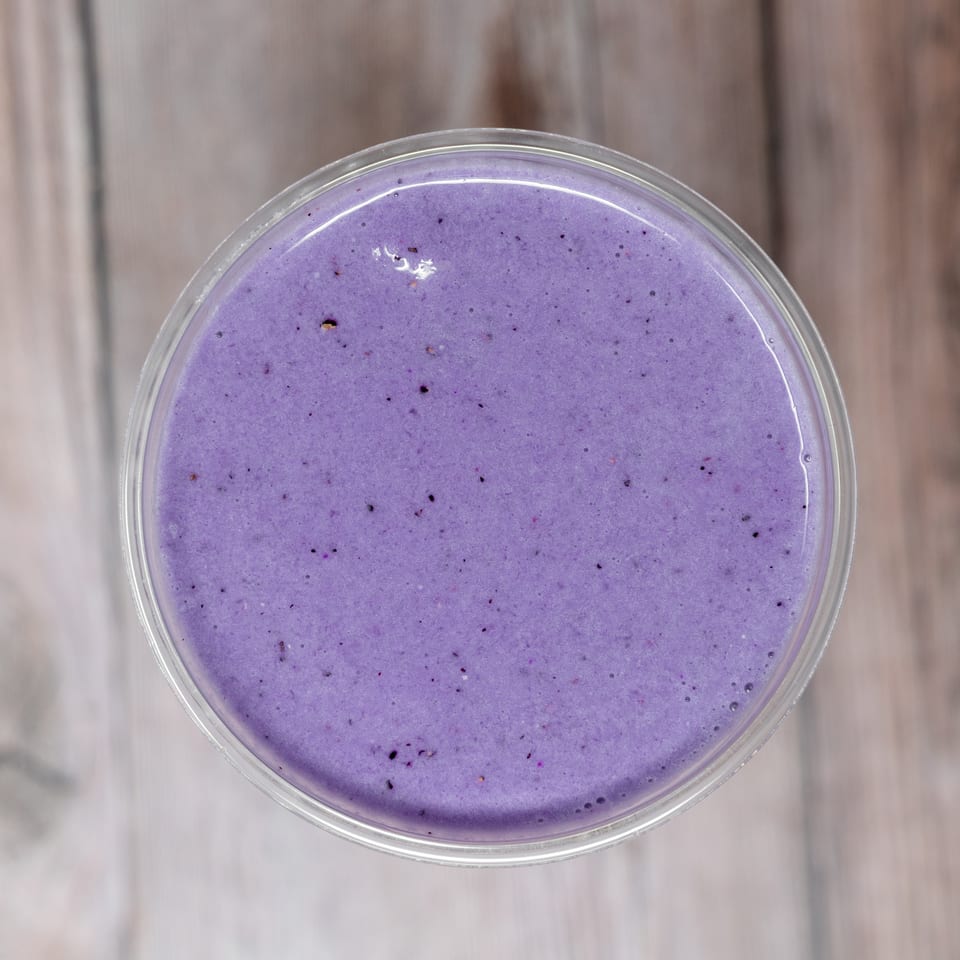 Top-down view of a purple berry smoothie in a clear glass on a light wooden table.