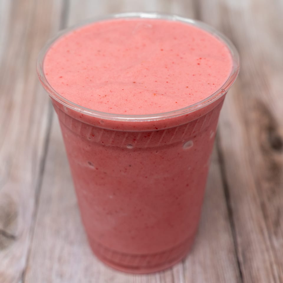 Pink strawberry smoothie in a clear plastic cup on a wooden table.