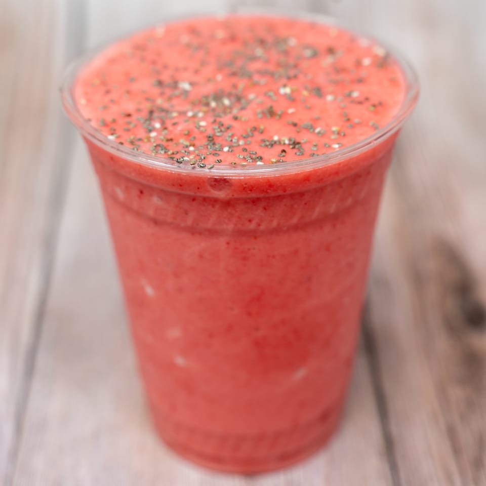 Red blended smoothie in a clear cup, topped with chia seeds and served on a wooden table.